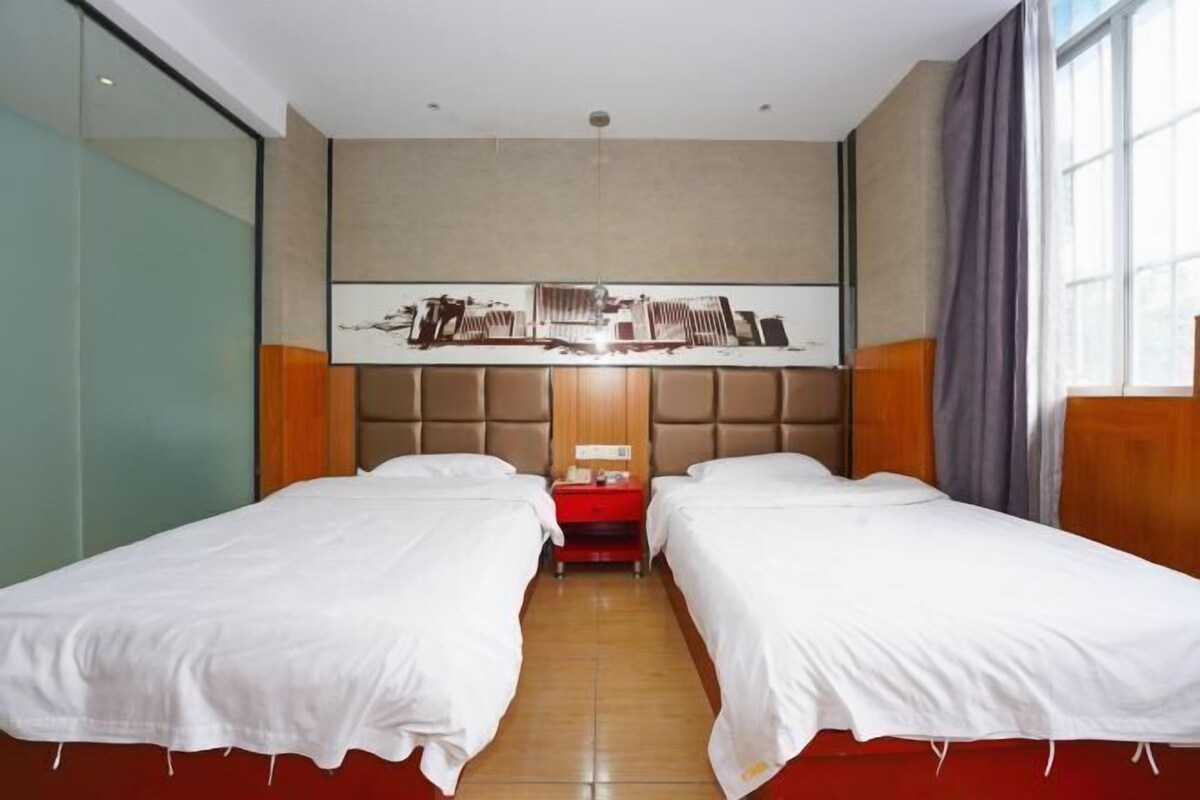Quarto de hotel com duas camas de solteiro, cabeceira de madeira, mesa de cabeceira vermelha e uma janela grande com cortinas.