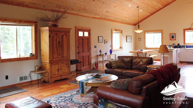 A spacious living room with a wooden ceiling, a brown leather sofa, a wooden cabinet, and a dining area with a table and chairs.