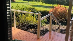 Eine Holzterrasse mit Geländer, die auf eine grüne Landschaft mit Bäumen und einem klaren Himmel blickt.