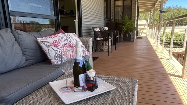Ein Balkon mit einer Korbsitzgruppe, einem Tisch, einer Weinflasche, Gläsern und einer Gartenzwerg-Figur.
