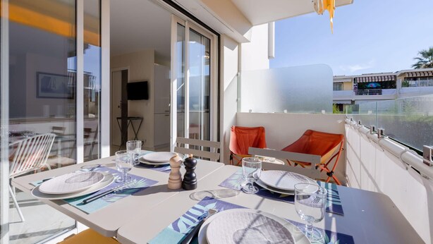 A dining area with a table set for four, a sliding glass door leading to an outdoor balcony, and a view of a building across the street. - Luxurious 2-room Apartment in Palm Beach (Cannes)