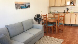 Un espacio habitable compacto con sofá gris, una mesa de comedor con sillas, un ventilador y una zona de cocina con armarios y suelo de baldosas.