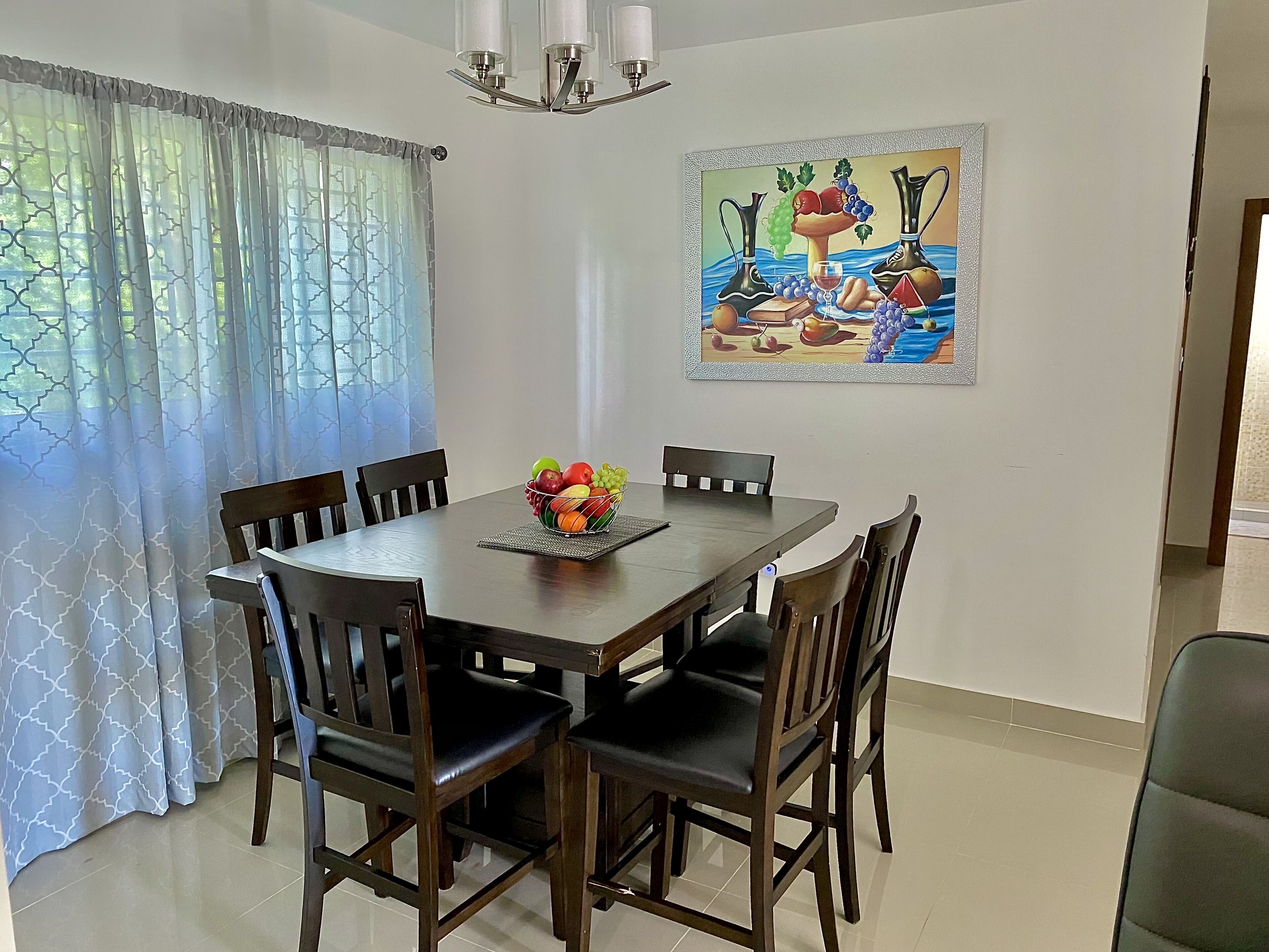 A dining area with a round table, six chairs, a fruit bowl, and a painting on the wall.