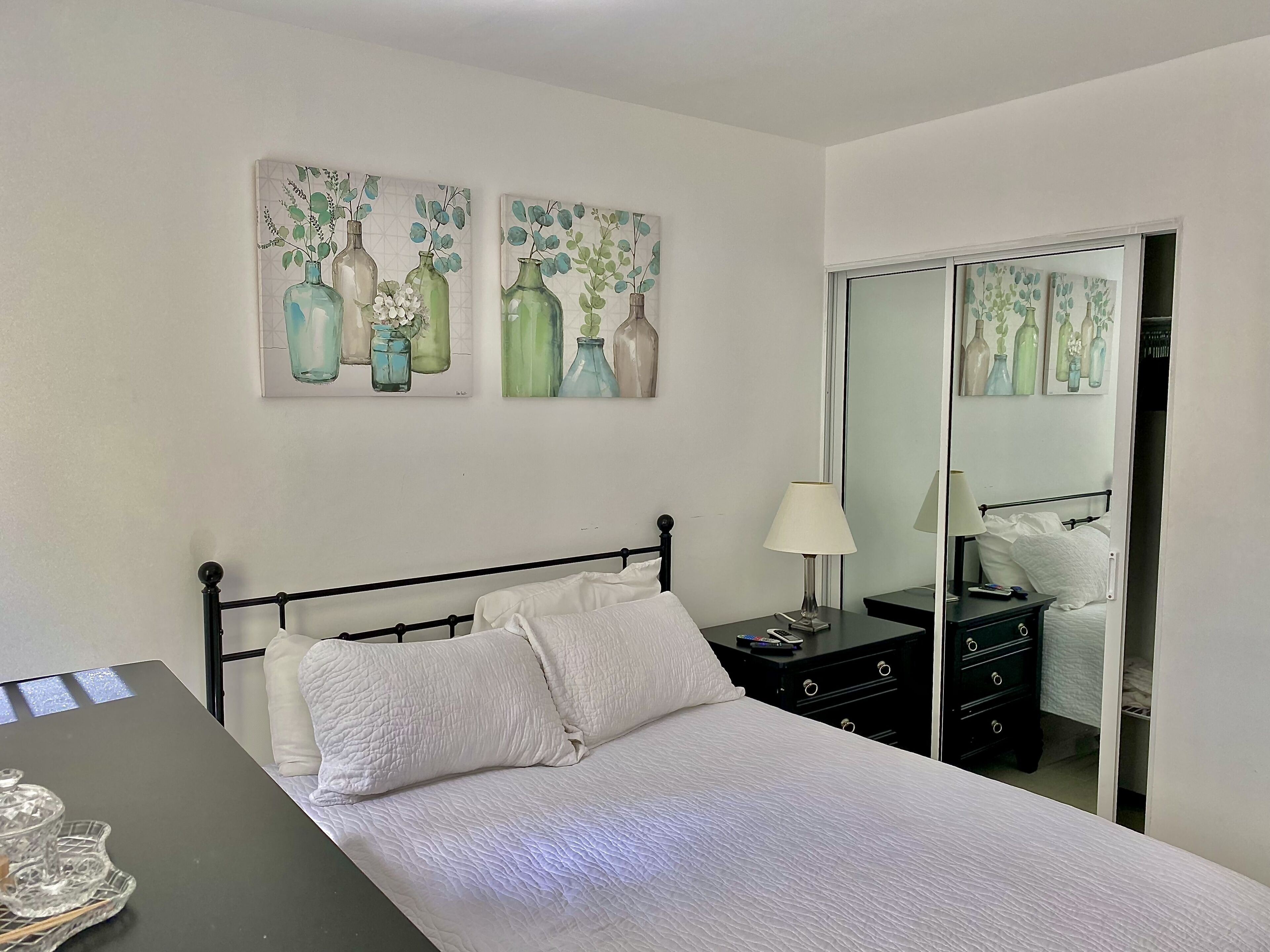 A neatly made bed with white bedding, a bedside table with a lamp, and a dresser with drawers. There are two wall-mounted paintings of vases with flowers.
