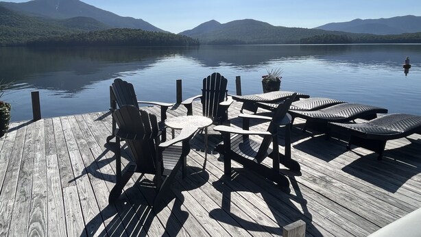 A wooden deck with Adirondack chairs and a table overlooking a calm lake with mountains in the background.