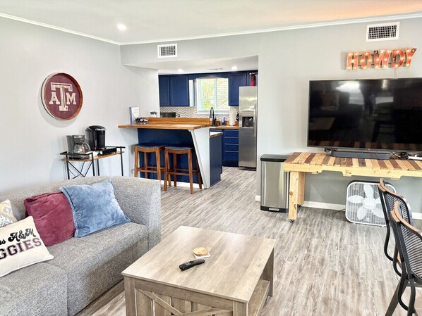 Ein modernes Wohnzimmer mit einem grauen Sofa, einem Holztisch und einem Fernseher. Im Hintergrund ist ein Küchenbereich mit blauen Schränken und einer Holzinsel erkennbar.