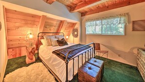 A bedroom with a wooden ceiling, a bed with a quilt, a bench, a dresser, and a window with a view of trees.