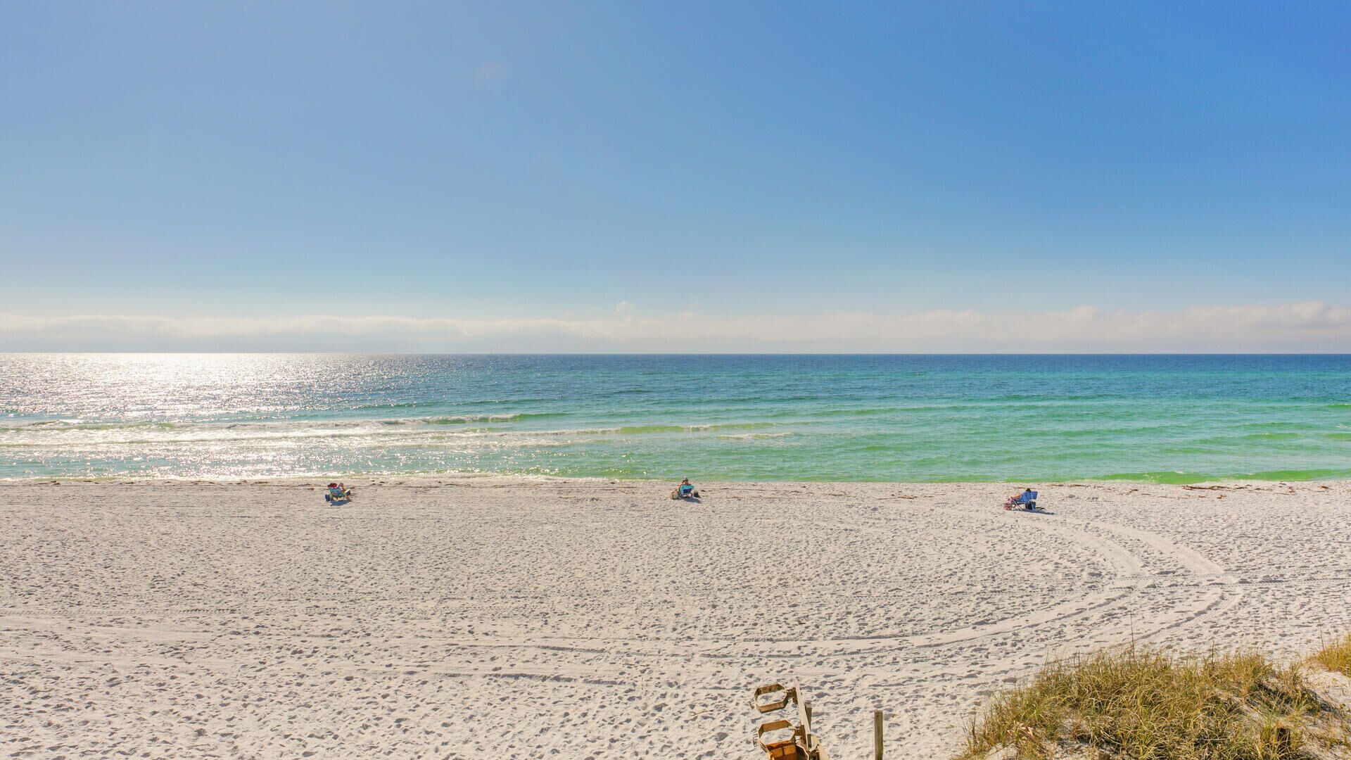 A wide sandy beach with clear blue water and a few people relaxing.