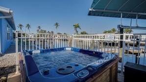 A hot tub with a wooden deck and a metal railing, surrounded by a pool area with palm trees and a clear sky. - 50’ of Dock for your boats. Hot tub, Boatlift, walk 2 restaurants Xtra parking.  (Matlacha)