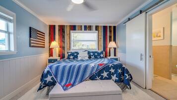 Une chambre à coucher comprenant un lit, des tables de chevet, un banc, une fenêtre avec des stores, un ventilateur de plafond et une porte.