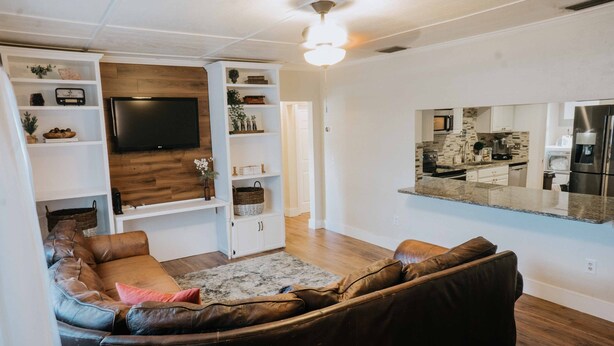 A modern living room with a brown leather sectional sofa, a flat-screen TV mounted on a wooden wall, and a kitchen area visible through an open doorway. - Charming Palm Beach Home Close to Beaches (Tequesta)