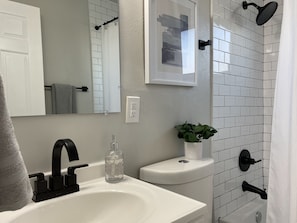 A bathroom with a white sink, black faucet, a mirror, a potted plant, and a shower area with white tiles.