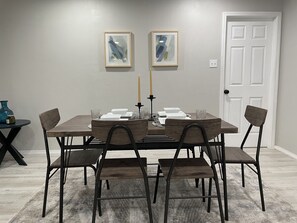 A dining room with a rectangular table set for four, surrounded by four chairs. There are two framed abstract paintings on the wall above the table.
