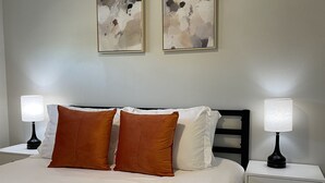 Una cama bien hecha con ropa de cama blanca y naranja, dos mesitas de noche con lámparas y arte abstracto en la pared.