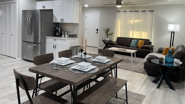 Una cocina moderna con armarios blancos, un refrigerador de acero inoxidable y un comedor con mesa y sillas de madera.