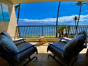 A balcony with two lounge chairs, a small table, and a view of the ocean and mountains.