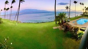 A pool area with a view of the ocean, surrounded by palm trees and a grassy lawn. - LUANA MOANA SIGNATURE OCEANFRONT MAUI-Ocean&Haleakala Views-FreeWiFi-A/C-Romance (Wailuku)