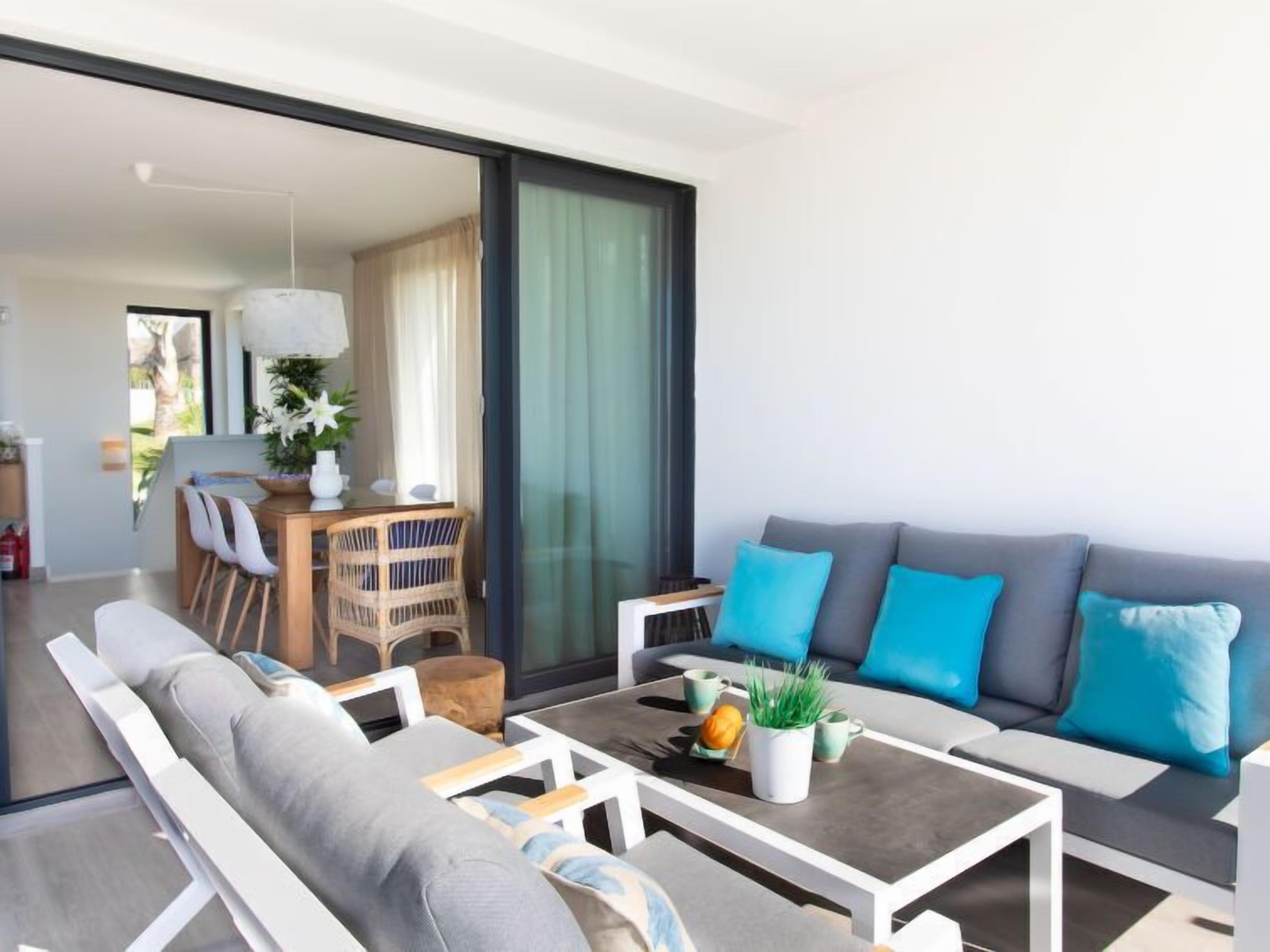 Ein modernes Wohnzimmer mit einer grauen Couch, weißen Sesseln und einem Couchtisch. Es gibt eine Essecke mit Tisch und Stühlen, sowie ein großes Fenster mit Vorhängen.