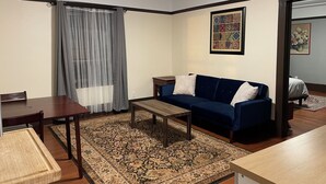 A living room with a blue sofa, a wooden coffee table, a dining table with chairs, a patterned rug, a large window with curtains, and a framed artwork on the wall. - Spacious 1-bedroom in Oakland with AC (Oakland)