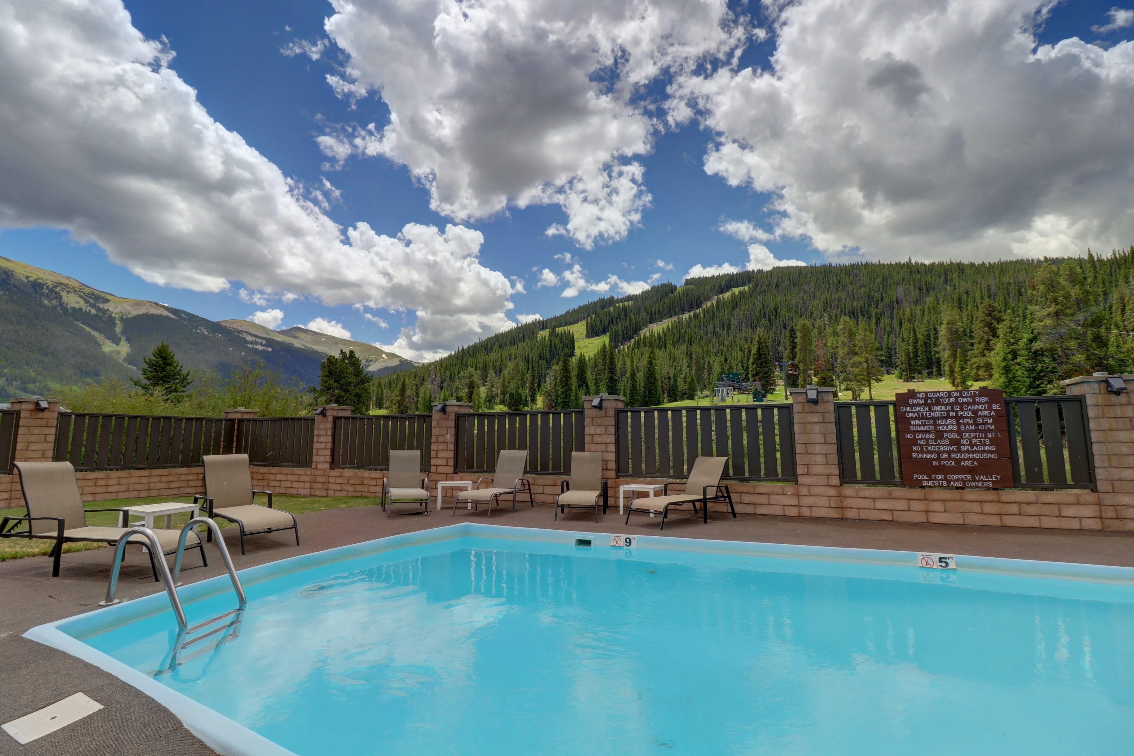 Una piscina con lettini e un cartello, circondata da un muro di mattoni e affacciata su una vista di montagne.