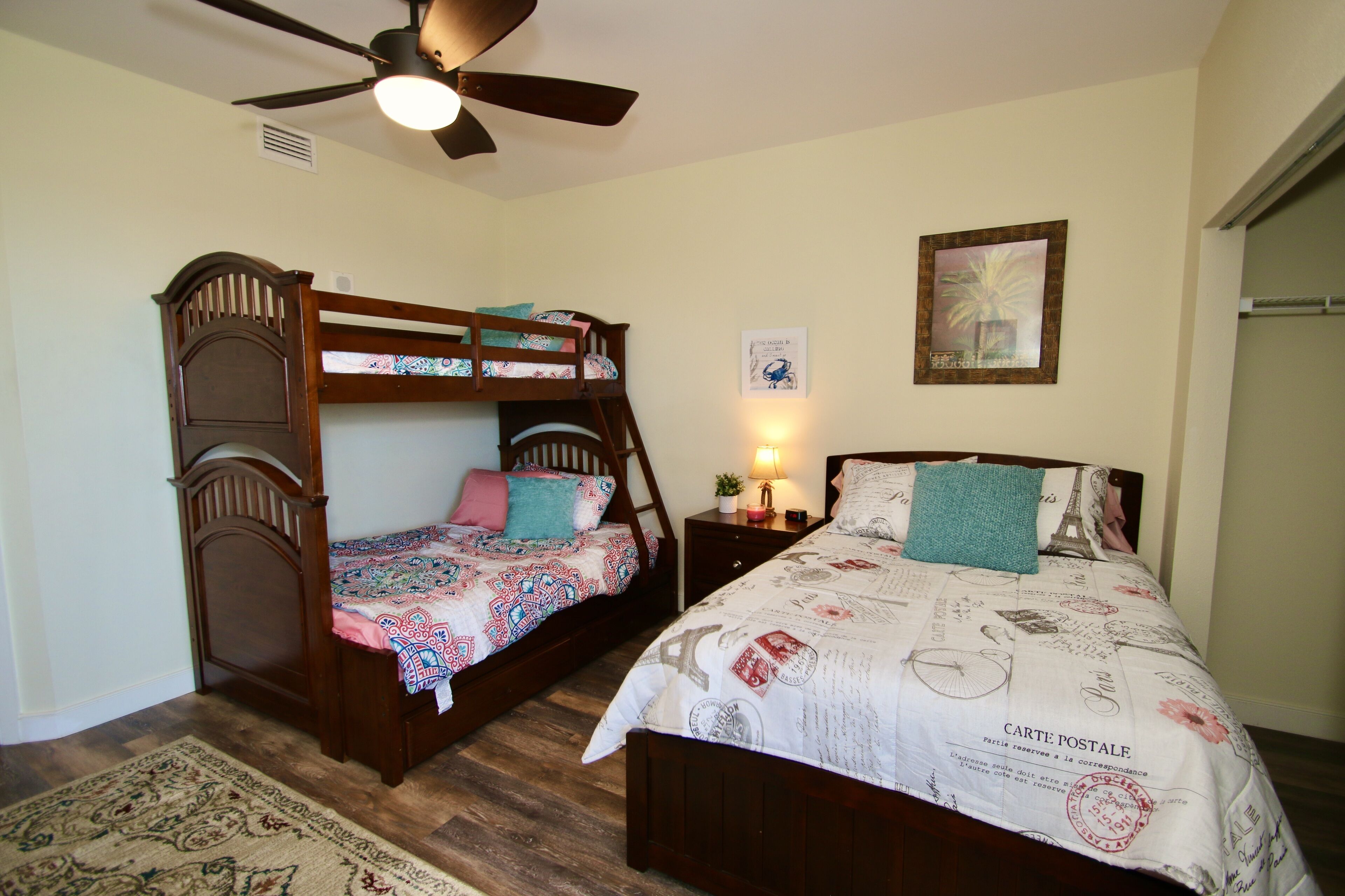 A bedroom with a bunk bed, a single bed, a ceiling fan, a nightstand with a lamp, and a framed picture on the wall.