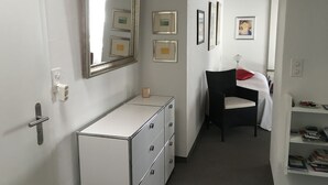 A hotel room with a white dresser, a mirror, framed pictures on the wall, a chair with a red pillow, and a bookshelf with books. - WEF Business Apartment (Davos)