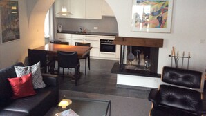 A living room with a fireplace, a dining area with a kitchen in the background, and a painting on the wall. - WEF Business Apartment (Davos)