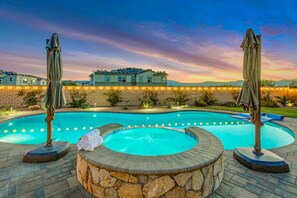 A swimming pool with a hot tub, surrounded by stone walls and two umbrellas. - Splash-Tastic Fun House (Indio)