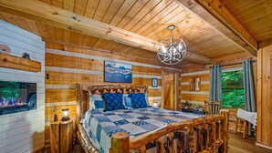 A wooden cabin bedroom with a large bed, wooden walls, a fireplace, and a view of greenery.