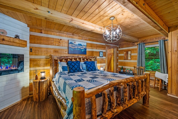 A wooden cabin bedroom with a large bed, wooden walls, a fireplace, and a view of greenery.