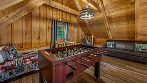 A cozy room with a wooden ceiling, a foosball table, a sofa, and a window with curtains.