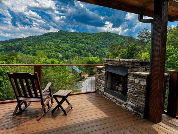 Une terrasse en bois avec une cheminée en pierre, un fauteuil à bascule et une vue sur une montagne boisée.