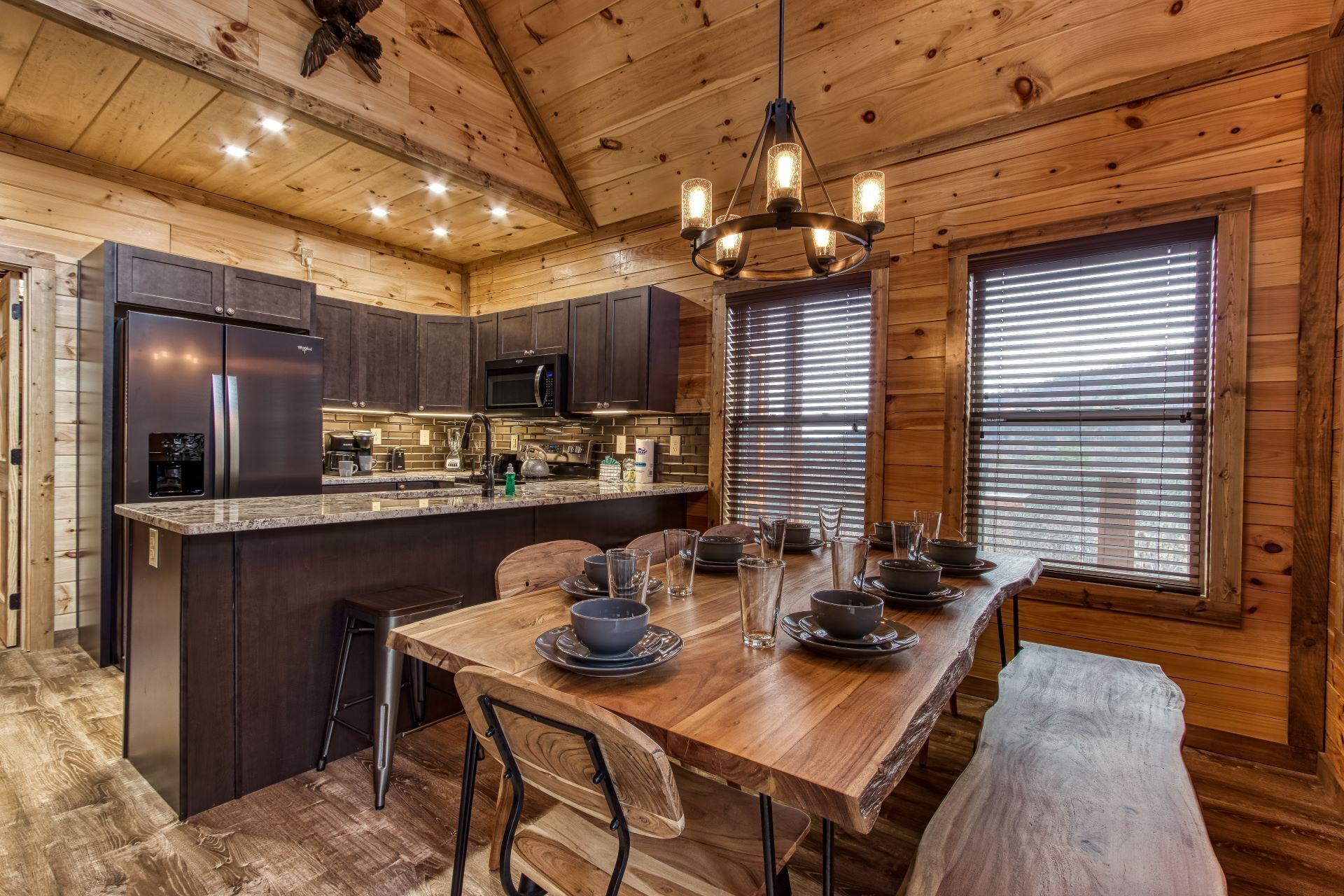 A wooden cabin interior with a kitchen, dining area, and a large window with blinds.