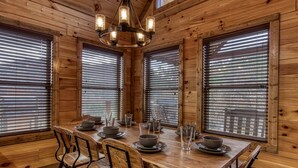 A wooden dining room with a large table set for four, flanked by large windows with blinds, and a chandelier above. - ERN805- BEARLY BELIEVE IT! - GREAT LOCATION! CLOSE TO TOWN AND ALL THE ACTION! (Pigeon Forge)