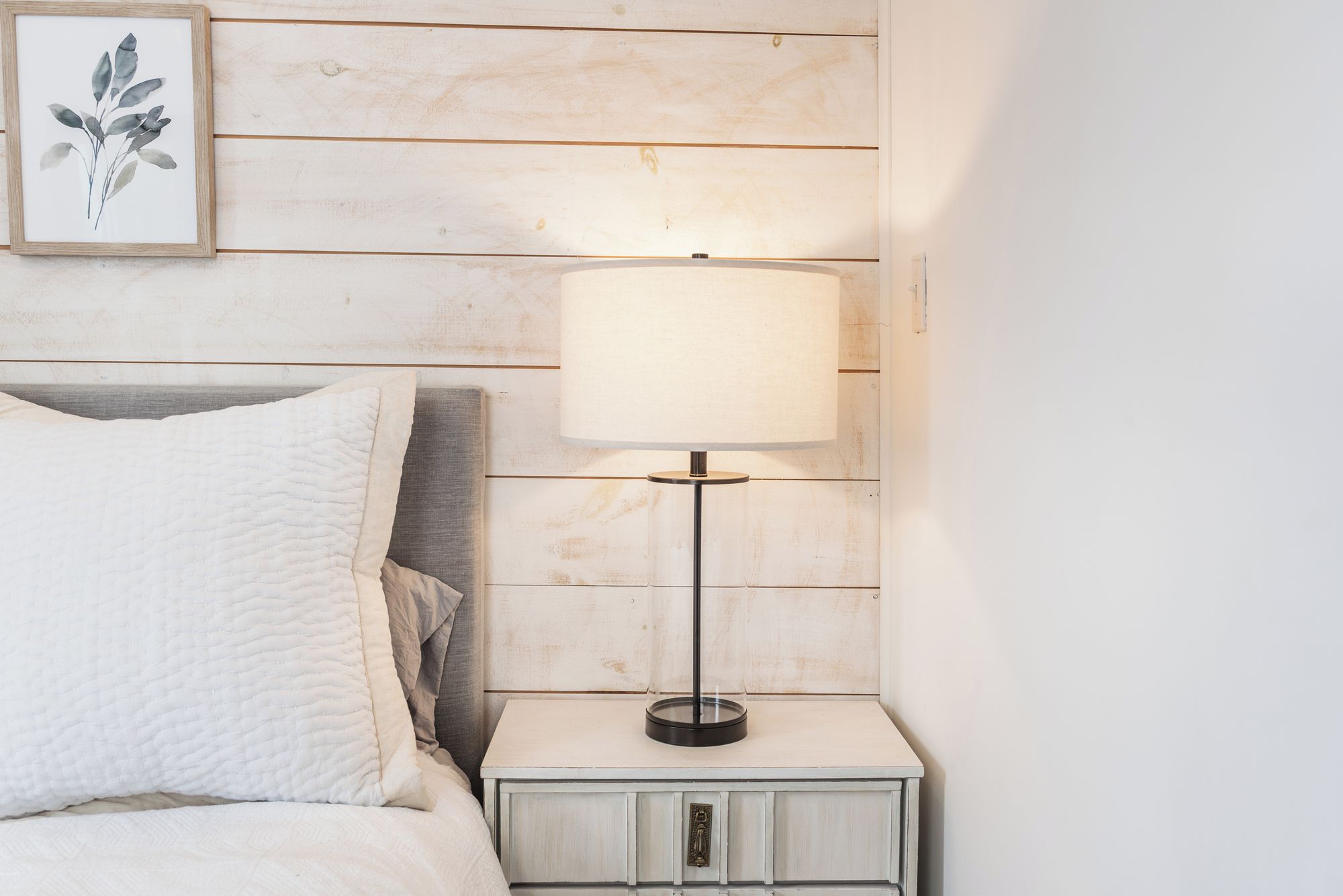 A neatly made bed with a white quilt, a bedside table with a lamp, and a framed botanical print on the wall.