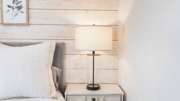A neatly made bed with a white quilt, a bedside table with a lamp, and a framed botanical print on the wall.