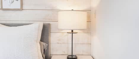 Een netjes opgemaakt bed met een witte quilt, een nachtkastje met een lamp, en een ingelijste botanische afbeelding aan de muur.