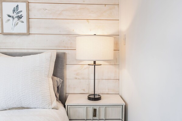 A neatly made bed with a white quilt, a bedside table with a lamp, and a framed botanical print on the wall.