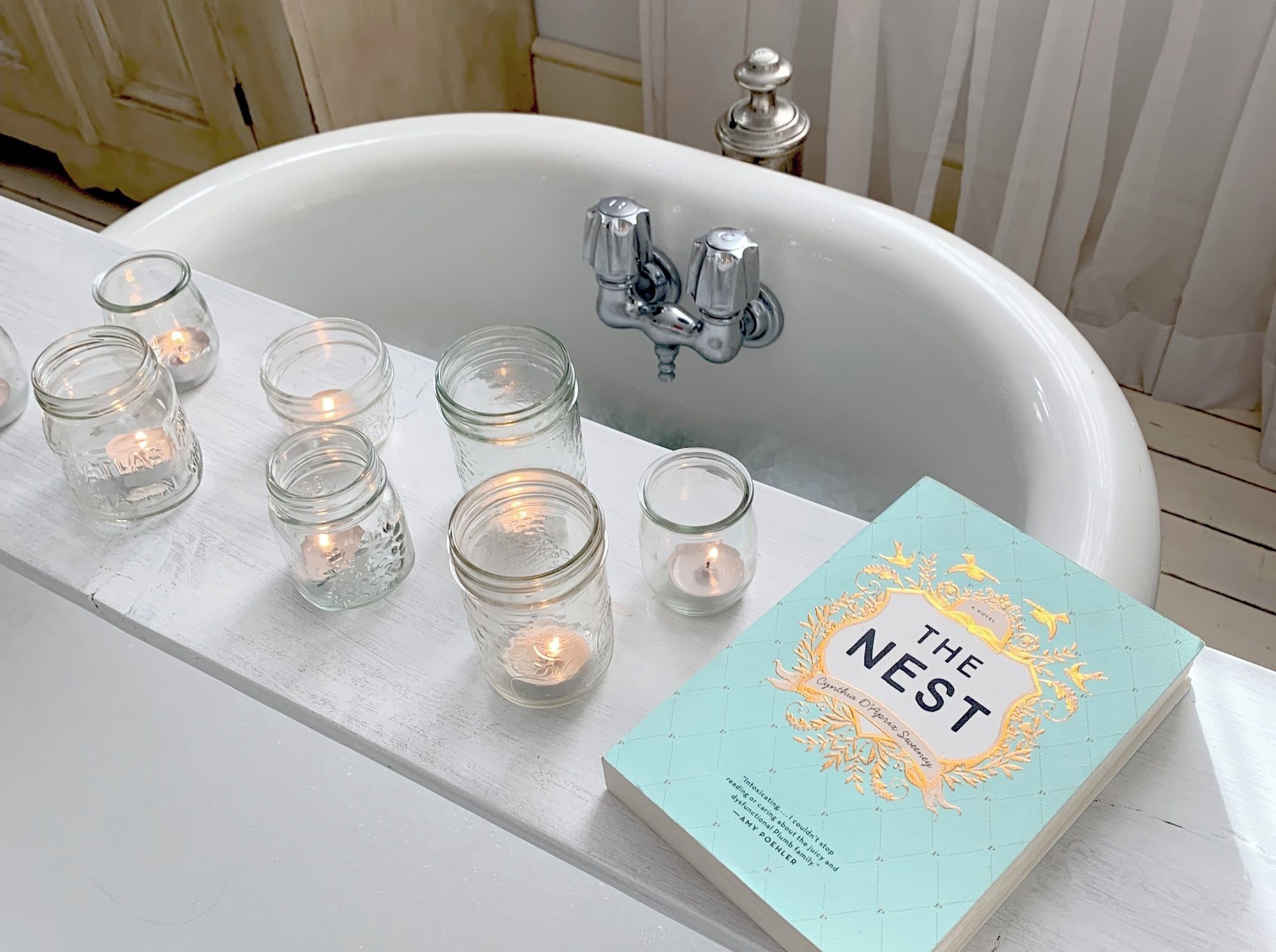 A white bathtub with a book titled 'The Nest' and several lit candles.
