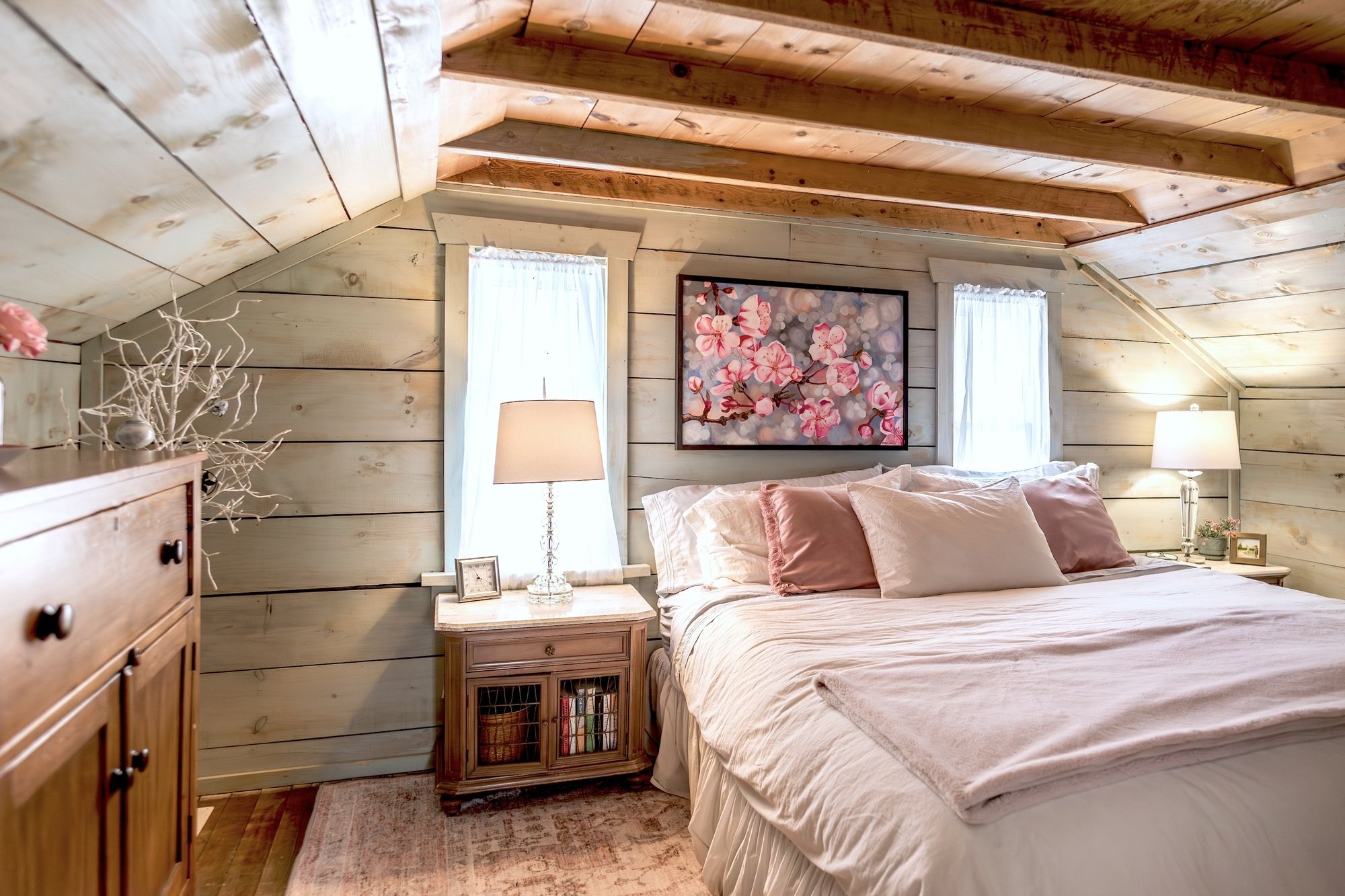 A bedroom with a sloped ceiling, wooden walls, a bed with white bedding, two bedside lamps, a nightstand with a lamp, a framed picture, and a vase with branches.