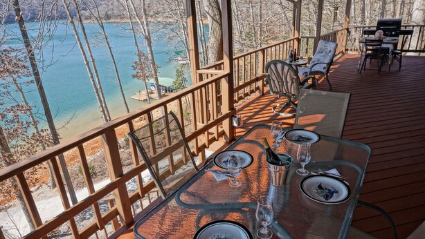 A dining area with a glass table set for two, overlooking a lake. - Norris Lakefront Retreat- Hot Tub, Firepit, Dock. Direct Lake Front. Sleeps 20 (LaFollette)