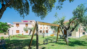 A playground with swings and a small table, surrounded by grass and trees, with a building in the background. - Cosy apartment for 6 people with WIFI, pool, A/C, terrace and pets allowed (Orosei)