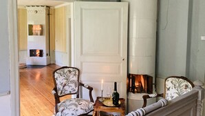 A room with a fireplace, two upholstered chairs, a small table with a candle and a bottle, and a mirror on the wall.