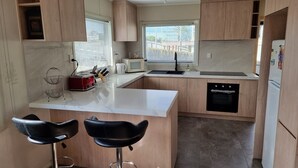 A modern kitchen with wooden cabinets, a white countertop, a microwave, a sink, a dishwasher, and two black bar stools.
