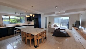 A modern kitchen with a central island, dining area with chairs, a sofa, and a TV. - Tide at Blueys Beach - Dog Friendly Beachfront (Blueys Beach)