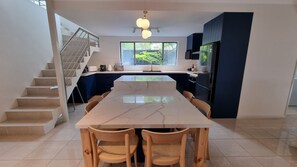 Una cocina moderna con una isla de mármol, sillas de madera, un refrigerador de acero inoxidable y una escalera que sube a otro nivel.