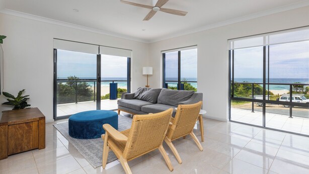 A spacious living room with a grey sofa, two wicker chairs, a blue ottoman, and a ceiling fan. Large windows offer a view of the ocean and a parked truck. - Tide at Blueys Beach - Dog Friendly Beachfront (Blueys Beach)