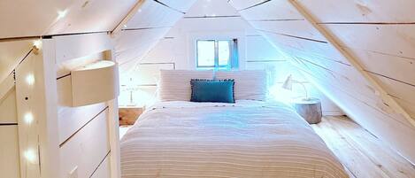 Un dormitorio con techo inclinado, una cama con ropa de cama a rayas, una ventana y una lámpara de noche.