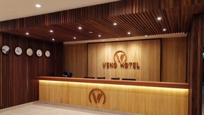 A hotel reception area with a wooden reception desk, a large logo, and clocks on the wall. - Veno Hotel (George Town)