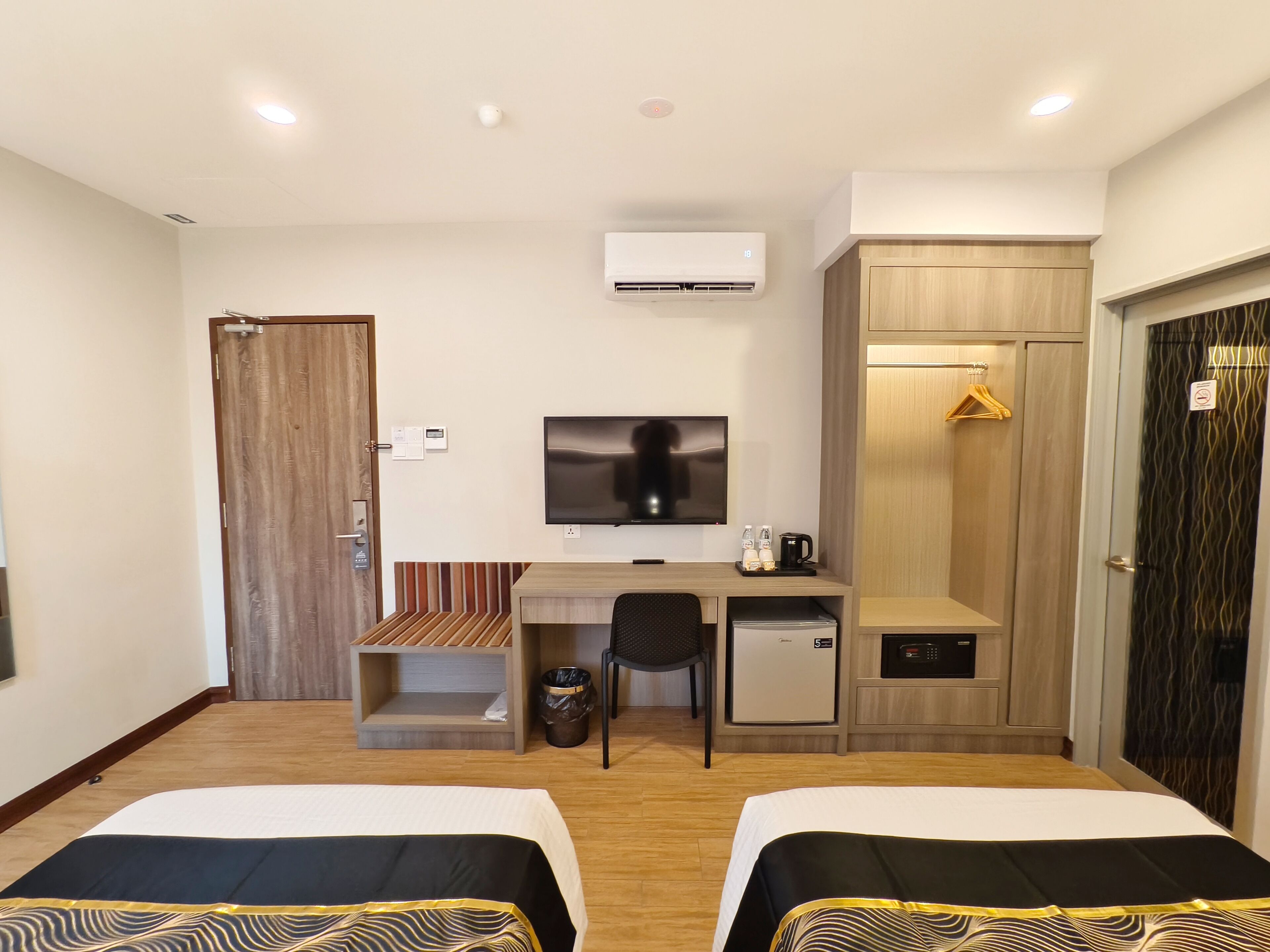 A hotel room with two beds, a desk with a chair, a television, a wardrobe, a small refrigerator, and an air conditioning unit.